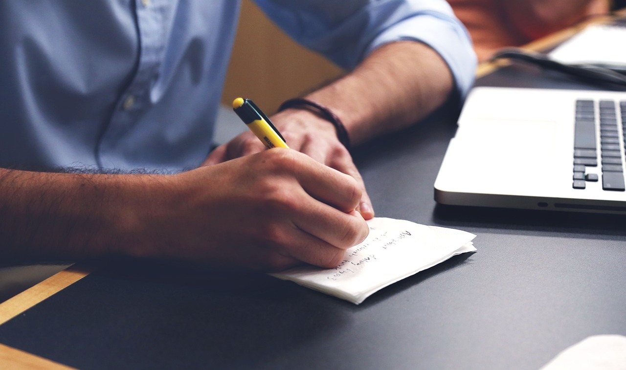 A person writing a note on a desk next to a laptop